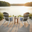 Two Kanyon Living Poly Folding Adirondack Chair Deluxe Driftwood Grey and White face a tranquil lake at sunset. A small table with snacks and drinks adds comfort to this cozy, coastal-inspired outdoor space surrounded by lush greenery.