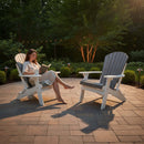 A woman in a light dress reads barefoot in a Kanyon Living Poly Folding Adirondack Chair Deluxe Driftwood Grey and White on a stone patio, beside another chair. Lush greenery surrounds them, with string lights overhead.