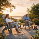 A relaxed couple enjoys sunset on a stone patio, drinks on a table, and their golden retriever nearby. They sit in Kanyon Living's Kanyon Poly Folding Adirondack Chairs in Driftwood Grey amid lush greenery by a serene lake.