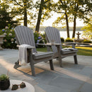 Two Kanyon Living Kanyon Poly Folding Adirondack Chairs in Driftwood Grey sit on a stone patio by a lake at sunset, surrounded by greenery and flowers. A coffee mug, book, and blanket rest on the chairs in this peaceful outdoor setting.