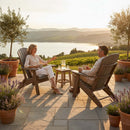 A woman and man relax on Kanyon Living's Kanyon Poly Folding Adirondack Chairs in Light Grey & Weathered Wood, seated on a stone patio with potted plants, enjoying sunset views of scenic hills, vineyards, and a lake.