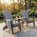 Two Kanyon Poly Folding Adirondack Chairs in Light Grey & Weathered Wood by Kanyon Living sit on a stone patio with a table and lemonade, one draped in a blanket, surrounded by hydrangeas and sunlit trees—ideal outdoor seating.