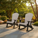 Two Kanyon Living Poly Folding Adirondack Chairs in Seashell & Black sit on a stone patio surrounded by greenery and flowers, providing comfortable outdoor seating. A drink is placed on the armrest between the chairs.