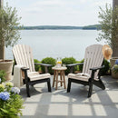 Two Kanyon Living Poly Folding Adirondack Chairs in Seashell & Black sit on a lakeside patio with a cozy blanket, hat, and lemonade on a small table, surrounded by lush plants and flowers.