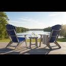 Two Kanyon Living Poly Folding Adirondack Chairs in Blue and Light Grey face a calm lake, separated by a small table with drinks and towel on a wooden deck surrounded by trees—the perfect outdoor setup.
