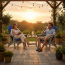 A man and woman relax in Kanyon Living’s Kanyon Poly Folding Adirondack Chairs in Blue and Light Grey on a patio at sunset, with potted plants, string lights, and snacks on a small table as they enjoy the scenic view.