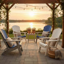 Four Kanyon Living Poly Folding Adirondack Chairs in Blue and Light Grey, with cushions, surround a table beneath a pergola lit by string lights. Potted plants and a cozy basket with blanket complete this beautiful lakefront sunset setting.