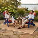 A man and woman relax with drinks on a patio, sitting in Kanyon Living's Kanyon Poly Folding Adirondack Chairs in red and blue. A small table with coffee, fruit, and books sits between them, with greenery and a lake in the background.