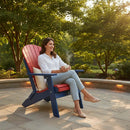 A woman in a white shirt and light jeans relaxes on a Kanyon Living Kanyon Poly Folding Adirondack Chair in red and blue, holding a steaming mug and smiling as she enjoys a sunny patio day surrounded by trees.