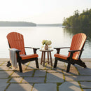 Two Kanyon Living Poly Folding Adirondack Chairs in Orange and Black sit on a stone patio by a calm lake. A small table with flowers, drinks, and a folded blanket rests between the chairs, creating an inviting outdoor scene.