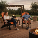 A woman reading and a man smiling relax on Kanyon Living Kanyon Poly Folding Adirondack Chairs in Orange and Black, made from recycled poly lumber, around a fire pit on a wooden deck at sunset—perfect outdoor furniture.