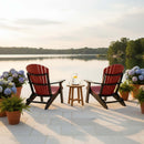 Two Kanyon Living Kanyon Poly Folding Adirondack Chairs in red and black face a calm lake at sunset, with a small table of lemonade between them. Potted hydrangeas and ferns line the patio, made serene by durable recycled poly lumber.