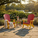 Two Kanyon Living Kanyon Poly Folding Adirondack Chairs in red and yellow sit on a stone patio with a small table between them. One chair has a blanket draped over it; flowers, plants, lush greenery, and a pond enhance the inviting outdoor scene.