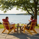 A woman and man sit in Kanyon Living's Kanyon Poly Folding Adirondack Chair in Red and Yellow by a lake, smiling at each other with a table between them, surrounded by green trees, water, and inviting outdoor seating.