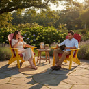 A woman and man relax outdoors on Kanyon Living Kanyon Poly Folding Adirondack Chairs in red and yellow, surrounded by greenery. She holds a drink, he reads a book, while a small table with a pitcher and plant sits between them.