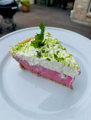 A slice of pie with a pink filling, made using Brushfire Farms Prickly Pear Simple Syrup, and topped with whipped cream, lime zest, and a sprig of mint, served on a white plate.