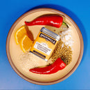 A plate with Spicewalla Citrus & Sesame Everything Seasoning, surrounded by red chili peppers, orange slices, sesame seeds, coriander seeds, and chopped onions on a blue background.