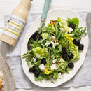 A fresh salad of leafy greens, blackberries, cheese, and nuts sits on a plate with a fork. Beside it is a bottle of Terrapin Ridge Farms Lemon Honey Vinaigrette by Terrapin Ridge Farms on a light wooden table.