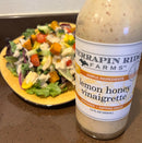 A bottle of Terrapin Ridge Farms Lemon Honey Vinaigrette stands in front of a vibrant mixed salad with lettuce, avocado, tomatoes, and veggies on a yellow plate.