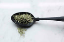 A matte black spoon holds a pile of World Spice Lemongrass by World Spice—dried, shredded lemongrass that's an essential Asian spice often used in herbal teas—resting on a white marble surface with a few stray shreds scattered below.
