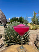 A bright pink margarita, crafted with Brushfire Farms Prickly Pear Simple Syrup, is garnished with a lime wedge and sits on a rock amid desert plants and cacti under a clear blue sky. The vibrant color and unique summery flavor come from South Texas prickly pear, courtesy of Brushfire Farms.