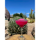A vibrant pink margarita made with Brushfire Farms Bulk Prickly Pear Simple Syrup and garnished with a lime wedge rests on a rock, surrounded by desert plants and a cactus garden under a clear blue sky.
