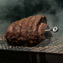 A large, seasoned roast is smoking on a grill with the FireBoard Pulse wireless smart meat thermometer from Fireboard inserted, surrounded by rising smoke against a dark background.