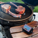 Two thick, bone-in steaks are cooking on a round charcoal grill outdoors, with a FireBoard Pulse wireless smart meat thermometer by Fireboard resting on a wooden side table next to the grill.