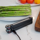 A FireBoard Pulse wireless smart meat thermometer by Fireboard, along with its charging cable, rests on a white counter next to a plate of fresh asparagus and a wooden cutting board.