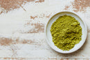A small white bowl filled with vibrant World Spice Culinary Matcha powder, made from finely milled green tea by World Spice, sits on a rustic, distressed white wooden table. The bowl is positioned on the right side of the frame, leaving space on the left.