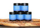 Five jars of World Spice Mayan Cocoa are stacked on a wooden board, surrounded by cinnamon sticks, peppercorns, and seeds. The jars feature blue labels and are arranged in a pyramid shape, evoking the rich tradition of chocolate and chile-infused drinking chocolate from World Spice.