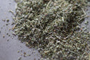 A close-up view of World Spice Mediterranean Mountain, a blend of dried green herbs by World Spice featuring indigenous mint family herbs, scattered on a gray surface.