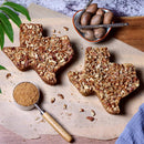 Savor authentic Texas flavor with the Texas Pecan Cakes Medium Texas Pecan Cake 2-Pack by Texas Pecan Cakes. Each cake, baked in the classic Lone Star shape and topped with generous chopped pecans, is presented on parchment and surrounded by whole pecans, brown sugar in a measuring cup, and a fresh green leaf—all beautifully arranged atop a wooden surface. This 2-pack serves 10-12, making it the perfect treat to share.