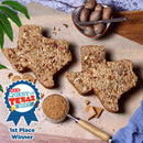 Two Medium Texas Pecan Cakes from Texas Pecan Cakes, each serving 10-12, sit atop a wooden board surrounded by whole pecans, a bowl of nutmeg with a grater, and a measuring cup filled with brown sugar. Representing genuine Texas flavor, the display features a blue ribbon label reading “H-E-B Quest for Texas Best, 1st Place Winner.”.