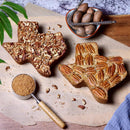 Two medium, Texas-shaped cakes from the Texas Pecan Cakes Medium Texas Pecan Cake 2-Pack by Texas Pecan Cakes rest on parchment paper—one adorned with chopped pecans, the other with pecan halves—capturing the true taste of Texas. Beside them, a bowl of whole pecans and a measuring cup of brown sugar highlight some of the delicious ingredients baked into each cake. Perfect for serving 10-12 people.