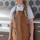Wearing a Messermeister Vintage Amber Leather Apron and a white chef’s coat, a person with a long braid stands before a stainless steel grill and hexagonal-patterned backsplash in a modern kitchen.