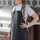 A woman in a white chef's jacket and a Messermeister Vintage Black Leather Apron stands by a stainless steel grill, hand on her hip, in a modern kitchen with a geometric tile backsplash.