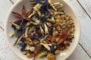 A white bowl sits on a rustic white wooden table, filled with World Spice Midnight Berry Tea by World Spice. The loose-leaf blend features blue butterfly pea flowers, yellow chamomile blossoms, cinnamon bark pieces, and two whole star anise pods—perfect for a soothing bedtime tea.