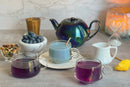 Three glass teacups filled with World Spice Midnight Berry Tea from World Spice, a black teapot, a milk pitcher, fresh blueberries, dried tea leaves, a lemon wedge, an hourglass, and a lit candle are arranged on a gray surface with a marble background.