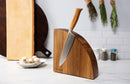 The Messermeister Oliva Elite Magnet Block Set – 6 Piece sits on a kitchen counter, its chef’s knife with an olive wood handle displayed in the curved wooden block amid chopping boards, spice jars, garlic cloves, and hanging herbs.