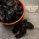 A bowl of smoky jalapeño peppers sits on a wooden surface with a few Rico Rico Chipotle Morita Dried Chiles – Smoky & Medium Heat Mexican Peppers beside it. Text in the corner reads, Fresh from the fields to your table, bringing you authentic Mexican flavor by Rico Rico.