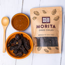 A bowl of chipotle Morita dried chiles, a wooden spoon, a bowl of red sauce, and a brown bag labeled Rico Rico Chipotle Morita Dried Chiles – Smoky & Medium Heat Mexican Peppers sit on a white wooden surface—perfect for adding the rich, smoky flavor of authentic Mexican jalapeño peppers to your favorite dishes from Rico Rico.