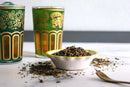 Two ornate glasses with gold and green patterns, filled with World Spice Moroccan Mint tea from World Spice, sit beside a small bowl of loose gunpowder tea leaves. Some leaves are scattered on a white surface, with a spoon nearby.