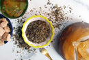 World Spice Moroccan Mint loose leaf tea in a small bowl, with scattered leaves, brown sugar cubes, a gold spoon, and a pitcher of iced tea on a white surface.