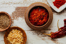 A wooden bowl filled with World Spice New Mexico Chili Powder by World Spice, surrounded by smaller bowls of dried herbs and garlic, a whole dried chili, and a plate with vibrant red chili powder on a rustic wooden surface—perfect for adding bold flavor to tacos or fajitas.