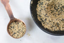 A copper measuring spoon holds World Spice Nigella Caesar—a coarse, tan and black seasoning featuring Worcestershire powder—and sits next to a black bowl filled with the same World Spice blend, all arranged on a white surface.
