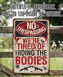 The No Trespassing Tired of Hiding The Bodies - Metal Sign by Lone Star Art features bullet holes, blood splatters, rifles, and a darkly humorous warning set against blooming flowers and greenery.