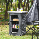 The DirectDeals Folding Camping Storage Cabinet with 3 Shelves and Carry Bag serves as a camp kitchen, storing drinks and snacks inside, while beer glasses and a can sit on top amid a tent and trees.