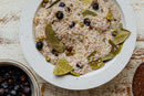 A white bowl filled with World Spice Northwest Forest Brine—a seasoned salt blend from World Spice featuring bay leaves, dried juniper berries, green herbs, and alderwood smoked salt—rests on a rustic wooden surface, perfect for brining meats.