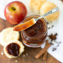 A jar of Old School Brand Apple Butter with a spoon on top, surrounded by biscuits, a red apple, cinnamon sticks, and spices on a wooden surface.